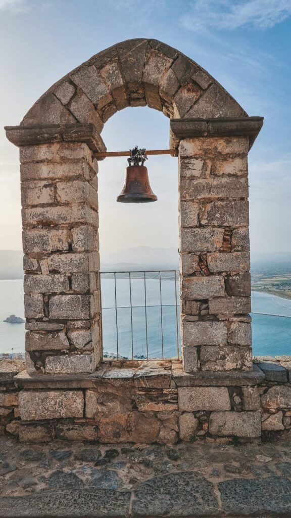 Sino antigo pendurado em arco de pedra na Fortaleza de Palamidi, com vista ao fundo de Nafplio
